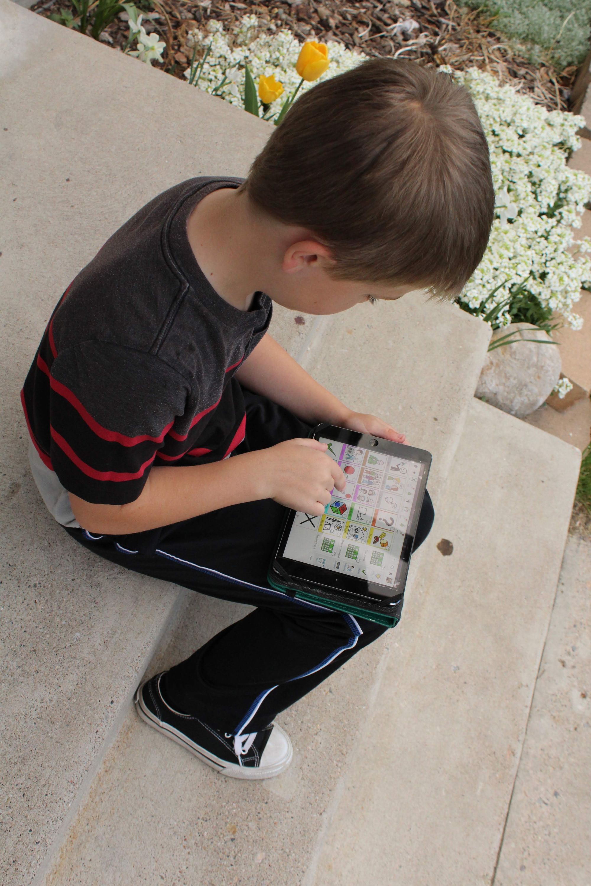 Young boy sits on cement steps with an AAC device in his lap as he presses button on the screen.