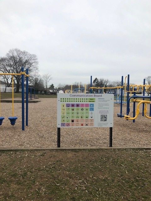 A blue and yellow playground is shown in the background.  Forground is a large, printed AAC speech board also displaying a scannable QR code.